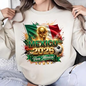 Puede incluir: Una sudadera de cuello redondo color crema con un estampado gráfico de la bandera de México, un trofeo dorado, un balón de fútbol y el texto "MEXICO 2026" en letras doradas, con una cinta verde debajo que dice "Viva México!" y cinco estrellas.