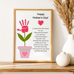 Könnte beinhalten: Ein gerahmter Druck zum Muttertag mit einem Handabdruck-Blumen-Design. Der rosa Handabdruck bildet die Blüte der Blume, mit grünen Blättern und einem rosa Topf. Der Text lautet "Happy Mother's Day!" und ein Gedicht.