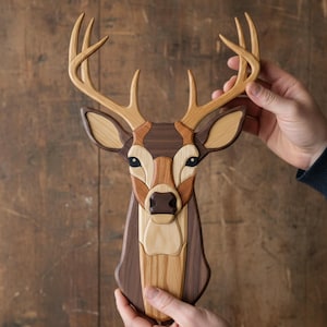 May include: A wooden deer head wall art piece. The deer head is made from various wood tones, including light and dark browns. The antlers are a lighter shade of brown. The deer's eyes are dark, and the design is detailed and artistic.
