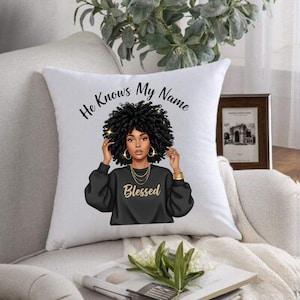 Könnte beinhalten: Weißes Zierkissen mit einer Illustration einer Frau mit lockigem schwarzem Haar, Goldschmuck und einem schwarzen Pullover mit dem Wort "Blessed". Das Kissen hat den Text "He Knows My Name" in Schwarz.
