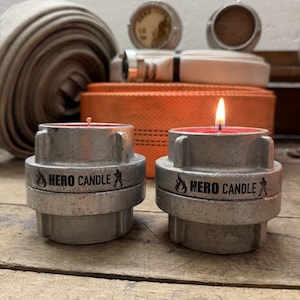 Könnte beinhalten: Zwei zylindrische Kerzenhalter aus Metall, gefertigt aus Feuerwehrschlauchkupplungen. Jeder Halter trägt den Schriftzug HERO CANDLE mit einem Flammen- und Feuerwehrman-Symbol. Eine Kerze ist unangezündet mit rosa Docht, die andere brennt.