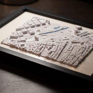 May include: A framed 3D relief map featuring a white architectural model of a city layout with raised buildings and streets. The map is set against a light background and enclosed within a simple black rectangular frame, creating a detailed display.