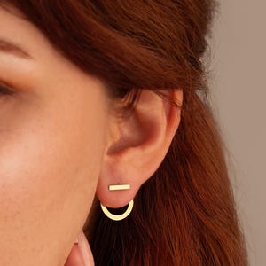 May include: A pair of gold earrings with a minimalist design. One earring is a small, thin bar, and the other is a half-circle with a bar extending from the top.