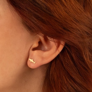 Puede incluir: Primer plano de la cara de una mujer, mostrando su oreja izquierda con un pequeño pendiente de tiburón de oro. Su cabello es largo y rojo, y está mirando hacia un lado.