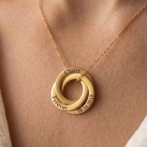 May include: A gold-colored necklace with three interlocking rings. The top ring is engraved with the name "Claire", the left ring says "Forever", and the right ring has the date "08.10.2019". The necklace is worn around a person's neck.