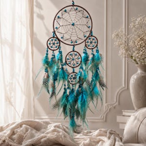 Op de afbeelding: Een bruine dromenvanger met een grote centrale ring en kleinere ringen eronder, versierd met turquoise kralen en veren. De dromenvanger hangt tegen een neutrale achtergrond en creëert een bohemien esthetiek.