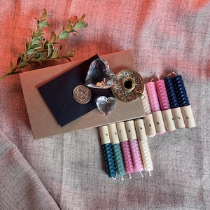 Puede incluir: Un surtido de artículos decorativos que incluyen velas, un candelabro de latón, un sobre negro con un sello de cera y adornos de cristal en forma de corazón. Las velas son de varios colores, incluyendo rosa, azul y blanco.