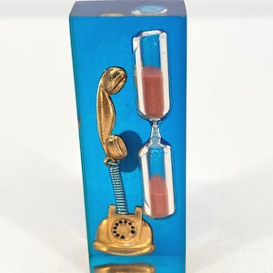 May include: A rectangular, blue resin paperweight featuring a gold-colored vintage telephone and a clear hourglass with red sand. The telephone has a coiled spring and a rotary dial. The hourglass is clear with red sand.