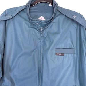 May include: A teal-colored jacket with a zip-up front and a stand-up collar. The jacket has shoulder epaulets and a small pocket on the chest. The text "WORLD TRAVEL CLUB" is visible on the pocket.