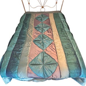 May include: A bedspread featuring a central vertical panel of iridescent mauve and teal diamond-shaped sections with embroidered details, flanked by wide vertical stripes of shimmering teal and blue fabric, draped over a metal bed frame.