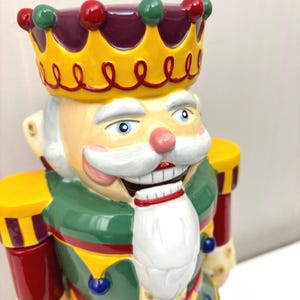 May include: A ceramic nutcracker figurine in the shape of a king. The figurine has a yellow crown with red and green accents, a white beard, and a green and red uniform. The nutcracker has a smiling face with blue eyes.