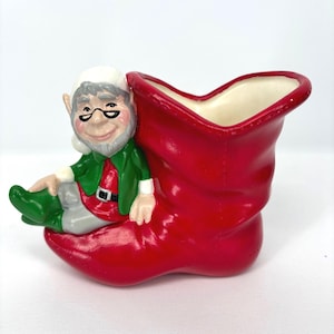 May include: A ceramic Christmas decoration featuring a red boot with a white interior. A smiling elf with a white hat, glasses, green jacket, and red shirt is seated on the boot. The elf has gray hair and a gray beard. The boot is a festive holiday decoration.