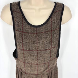 May include: A sleeveless brown dress featuring a textured plaid pattern with thin red grid lines. The garment has a wide scoop neckline and armholes finished with solid black trim, creating a structured look on the torso and bodice.