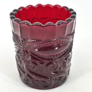May include: A ruby red glass tumbler with a scalloped rim and ornate, embossed detailing. The design features swirling patterns and small, raised dots. The glass has a vintage aesthetic and is likely a decorative item.