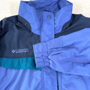 May include: A multi-colored Columbia Sportswear Company jacket. The jacket features a high collar and a button closure. The color scheme includes shades of blue, teal, and black. The Columbia logo is visible on the left chest area.