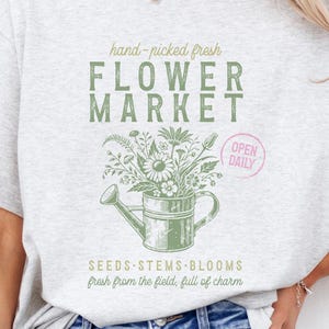 Peut inclure: Un t-shirt gris avec un graphique d'un arrosoir rempli de fleurs et le texte "hand-picked fresh FLOWER MARKET OPEN DAILY SEEDS STEMS BLOOMS fresh from the field, full of charm"