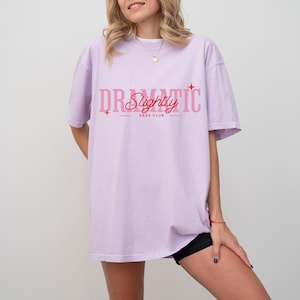 May include: A light purple t-shirt with the words "DR Slightly TIC" in pink, with "SASS CLUB" underneath. The shirt is a relaxed fit and has short sleeves.