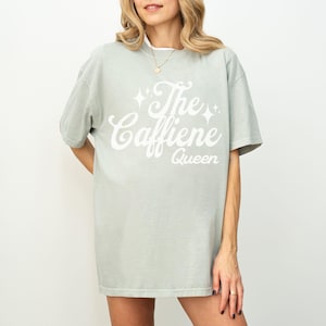 May include: A light green t-shirt with the text "The Caffeine Queen" in white script. The shirt has short sleeves and a relaxed fit. The text is embellished with starburst designs.
