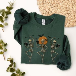 May include: Dark green sweatshirt featuring a Halloween-themed design. The graphic depicts three dancing skeletons holding books, with a large orange circle in the background, bats, and a crescent moon. The sweatshirt is made of a soft material.