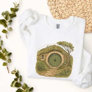 May include: White sweatshirt with a watercolour illustration of a hobbit hole. The design features a green door, stone path, and the text "Courage is found in unlikely places." Includes a small window and a tree.