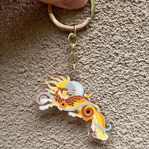 Puede incluir: Un llavero dorado con un dise&ntilde;o de lobo estilizado con un motivo de sol y luna. El lobo est&aacute; representado en un esquema de color amarillo y marr&oacute;n con un fondo blanco.