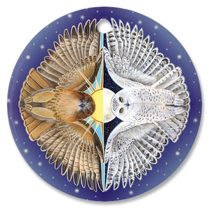 May include: A round, blue ornament with a white snowy owl and a brown hawk with outstretched wings. The owl is on the left side of the ornament and the hawk is on the right. The owl is facing the left and the hawk is facing the right. The background is a dark blue with white stars.