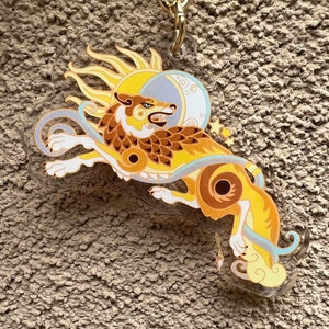 Puede incluir: Un lobo amarillo y marr&oacute;n con un dise&ntilde;o de sol y luna, en un llavero de acr&iacute;lico transparente. El lobo est&aacute; estilizado con un sol detr&aacute;s de &eacute;l y una luna creciente por encima.