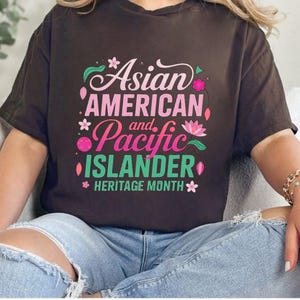 Peut inclure: T-shirt marron avec l'inscription "Asian American and Pacific Islander Heritage Month" en rose, vert et blanc, ornée de motifs floraux. Le vêtement est porté avec un jean bleu. Le design est attrayant.