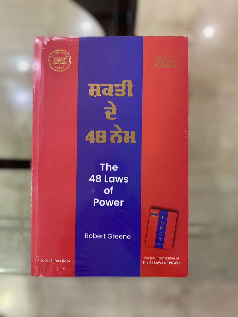 以下が含まれることがあります： ロバート・グリーン著「The 48 Laws of Power」という赤と青の表紙の本。この本はオリジナル作品のパンジャブ語訳です。本の表紙には「ਸ਼ਕਤੀ रे 48 तेभ」という金色の文字が書かれています。
