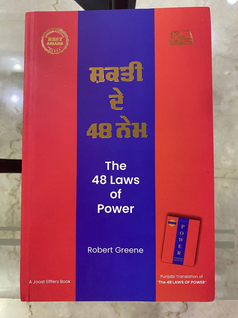 以下が含まれることがあります： ロバート・グリーン著「The 48 Laws of Power」というタイトルの赤と青の表紙の本。この本は、英語版のオリジナルバージョンからのパンジャブ語訳です。本の表紙には、「ベストセラー」と書かれた金色のシールがあります。