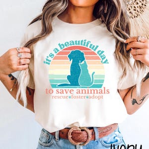 May include: An ivory t-shirt featuring a graphic of a dog and cat silhouette against a multi-colored sunset circle. The text reads: it's a beautiful day to save animals, rescue, foster, adopt. The design uses teal, orange, and yellow colors.
