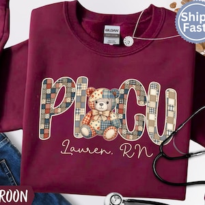 May include: Maroon sweatshirt with the letters "PICU" in a patchwork design, featuring a teddy bear. The name "Lauren, RN" is written below. A stethoscope and the word "MAROON" are also visible.