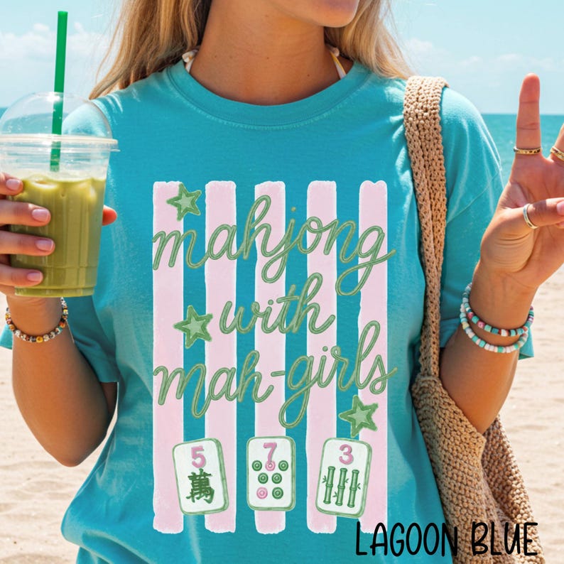 Op de afbeelding: Een turquoise t-shirt met een afbeelding van verticale roze strepen en de groene tekst "mahjong with mah-girls". Het ontwerp bevat drie groene sterren en drie mahjongtegels met de cijfers 5, 7 en 3 in groene inkt.