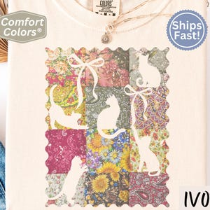 May include: Ivory-colored t-shirt featuring a patchwork design with floral patterns and white cat silhouettes. The shirt has the words "Comfort Colors" and "Ivory" printed on it. A necklace is visible.