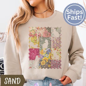 May include: Sand-colored sweatshirt featuring a patchwork design of floral patterns and cat silhouettes. The design includes various colors like pink, yellow, and green. The sweatshirt has long sleeves and a crew neck.