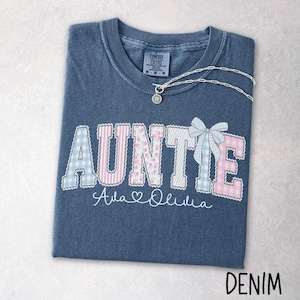 May include: A denim blue t-shirt with the word "AUNTIE" in a patchwork design, featuring light blue gingham, pink stripes, and floral patterns. A silver necklace with a pendant rests on the shirt. The names "Ava & Olivia" are written below.