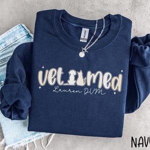 May include: A navy blue crewneck sweatshirt featuring the text "vet med" with a dog and cat silhouette integrated into the lettering, along with the name "Lauren DVM" in script. The design includes small star accents and a light plaid pattern.