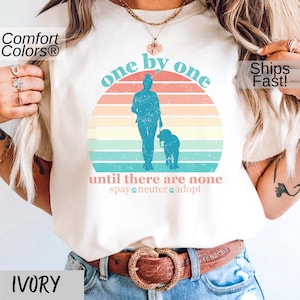 May include: A white t-shirt with a vintage-style graphic of a person walking a dog with the text "one by one until there are none spay neuter adopt".