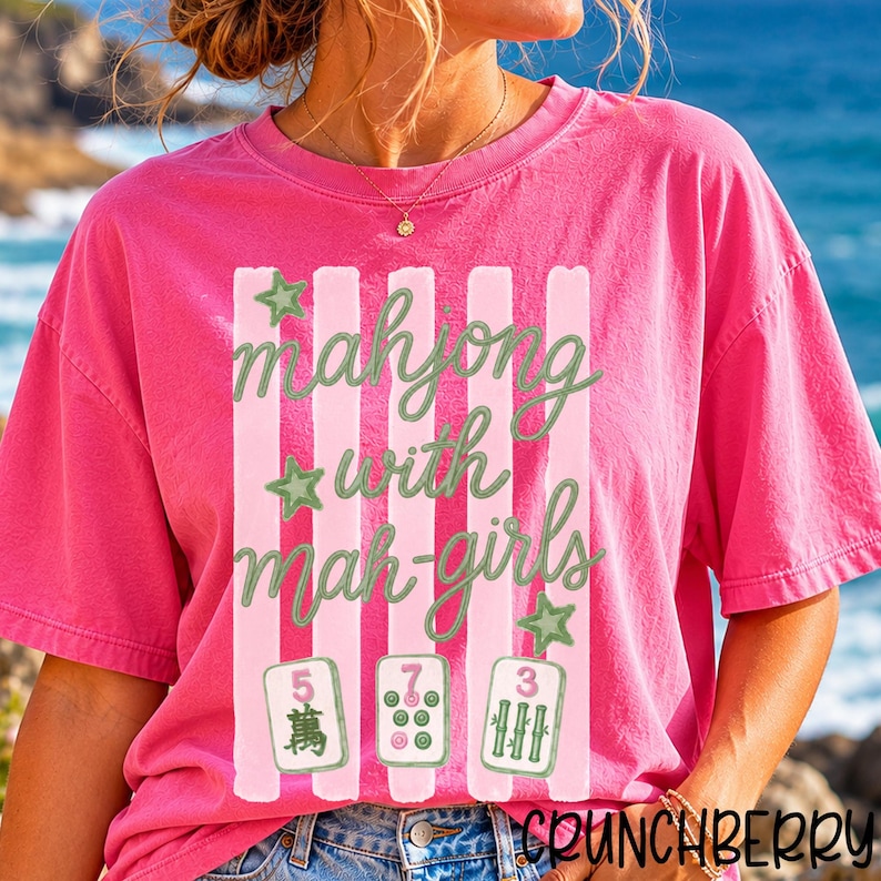 Op de afbeelding: Een felroze T-shirt met korte mouwen en een afbeelding van verticale lichtroze strepen, groene sterren en de tekst "mahjong with mah-girls" in groen schrift. Onder de tekst staan drie mahjongtegels met de cijfers 5, 7 en 3.
