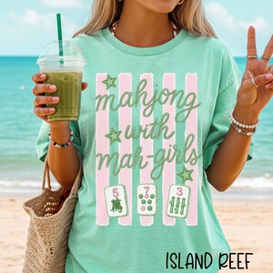 Op de afbeelding: Een mintgroen t-shirt met een afbeelding van verticale roze strepen, groene sterren en de tekst "mahjong with mah-girls" in een cursief lettertype. Onder de tekst staan drie mahjongtegels met de cijfers 5, 7 en 3 in het groen en roze.