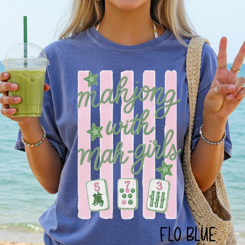 Op de afbeelding: Een blauw t-shirt met een afbeelding van verticale roze strepen, groene sterren en de tekst "mahjong with mah-girls" in groen schrift. Onder de tekst staan drie mahjongtegels met de cijfers 5, 7 en 3 en bijbehorende symbolen.