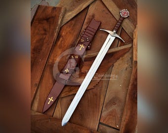 May include: A steel longsword with a silver-toned crossguard and a brown leather-wrapped grip featuring a circular pommel with a red cross emblem. The matching brown leather scabbard displays gold-colored cross accents along its length.