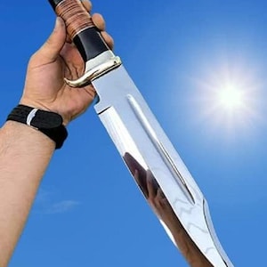 Könnte beinhalten: Ein großes, poliertes Messer aus Edelstahl mit gebogener Klinge. Der Griff ist schwarz und braun mit einem Messingansatz und Knauf. Das Messer wird vor einem hellblauen Himmel gehalten.