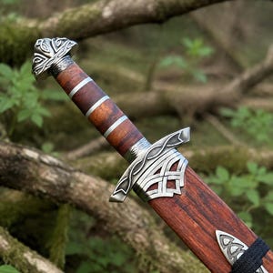May include: A sword with a dark brown wooden handle featuring silver metal bands and an ornate silver pommel. The crossguard has intricate silver metal knotwork detailing. The wooden scabbard includes a silver metal accent and a black cord wrap.