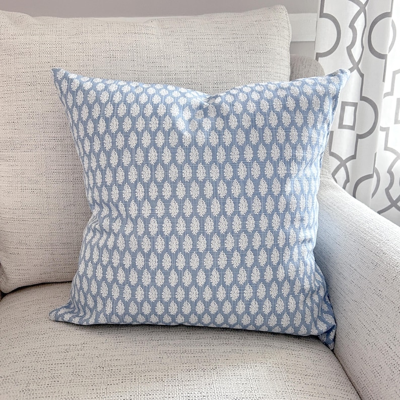 K&ouml;nnte beinhalten: Ein quadratisches Zierkissen mit hellblauem Hintergrund und einem wei&szlig;en Tropfenmuster. Das Kissen liegt auf einem hellfarbigen, strukturierten Sofa. Es ist ein Einrichtungsgegenstand.