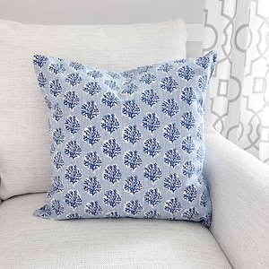 K&ouml;nnte beinhalten: Ein quadratisches Zierkissen mit hellblauem Hintergrund und einem sich wiederholenden Muster aus dunkelblauen korallenartigen Formen. Das Kissen liegt auf einem wei&szlig;en strukturierten Sofa, mit einem wei&szlig;en Kissen links.