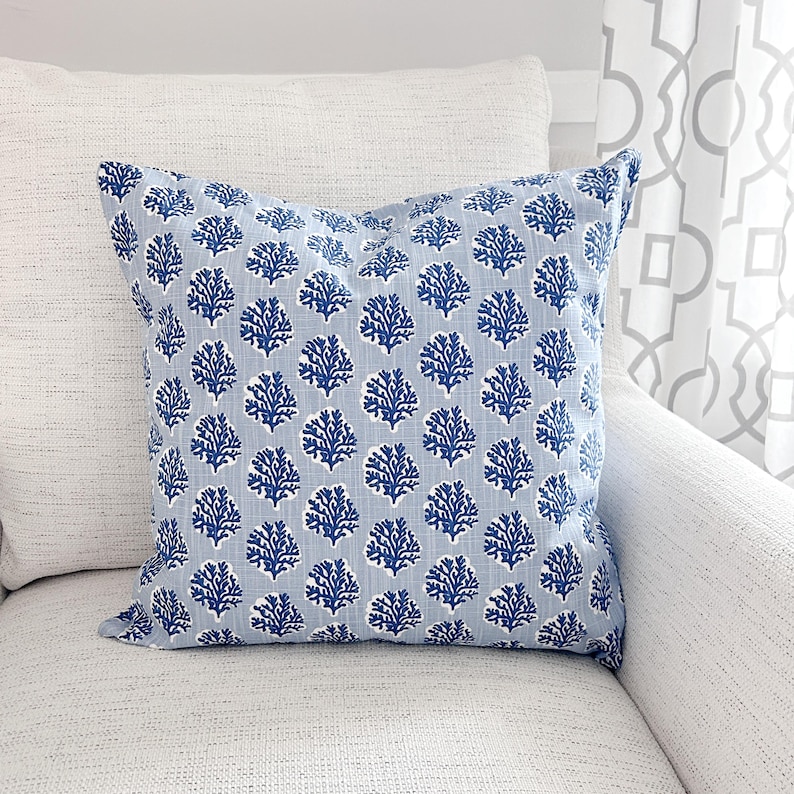 K&ouml;nnte beinhalten: Ein quadratisches Zierkissen mit hellblauem Hintergrund und einem sich wiederholenden Muster aus dunkelblauen korallenartigen Formen. Das Kissen liegt auf einem wei&szlig;en strukturierten Sofa, mit einem wei&szlig;en Kissen links.