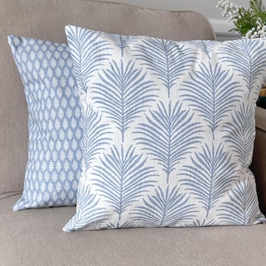 K&ouml;nnte beinhalten: Zwei dekorative Kissen mit blau-wei&szlig;en Mustern. Eines zeigt ein sich wiederholendes Blattdesign, das andere ein geometrisches Muster. Die Kissen liegen auf einem neutralfarbenen Sofa und verleihen dem Raum einen Hauch von Farbe und Textur.