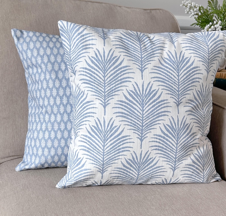 K&ouml;nnte beinhalten: Zwei dekorative Kissen mit blau-wei&szlig;en Mustern. Eines zeigt ein sich wiederholendes Blattdesign, das andere ein geometrisches Muster. Die Kissen liegen auf einem neutralfarbenen Sofa und verleihen dem Raum einen Hauch von Farbe und Textur.