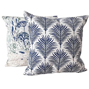 K&ouml;nnte beinhalten: Zwei quadratische Zierkissen. Eines hat einen wei&szlig;en Hintergrund mit einem navyblauen Palmenblattmuster. Das andere hat einen wei&szlig;en Hintergrund mit einem blau-gr&uuml;nen botanischen Design. Ideal f&uuml;r die Dekoration.